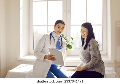 Portrait of a young doctor wearing stethoscope holding report file with appointment and giving consultation a smiling woman sitting on the couch in office during medical examination in clinic. - Powered by Shutterstock