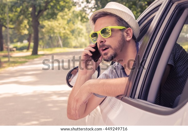Portrait of young\
driver with hat and sunglasses talking on smartphone from the car.\
Vacation and travel\
concepts.