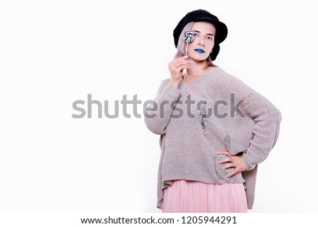 Portrait of young emotional attractive woman in black cap, torn sweater, with blue lips and pink hair, she is holding black and white lollipop, isolated on white background. 
