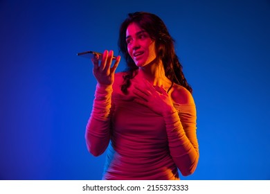 Portrait Of Young Emotional Girl, Student With Long Dark Hair Using Gadget Isolated On Dark Blue Background In Neon Light. Concept Of Emotions, Facial Expression, Youth, Aspiration, Sales.