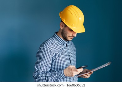 Portrait of young engineer or building contractor wearing yellow protective helmet and shirt standing in front of blue wall background holding smart phone and clipboard reading messages or report