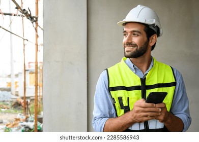 portrait of young engineer in vest with white helmet standing on construction site, smiling and holding smartphone for worker, internet, social media - Powered by Shutterstock