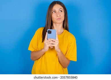 Portrait Of Young European Brunette Woman Wearing Yellow T-shirt On Blue Background With Dreamy Look, Thinking While Holding Smartphone. Tries To Write Up A Message.