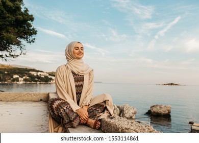 portrait of young European Muslim women with hijab sitting on the stone beach with sea in the background. She is happy and relaxed. - Powered by Shutterstock