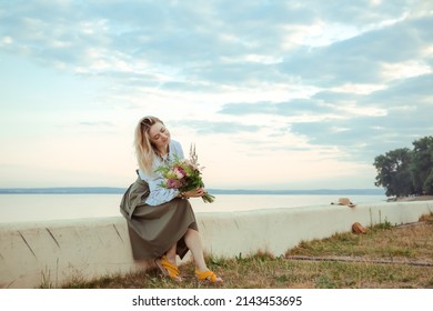 Portrait Of Young European Woman Sitting On The Curb With Bouquet Of Flowers. Beautiful Girl. Relaxing Morning Date Walk. Fashion. Natural Sea Landscape Background. International Woman Or Mother Day.