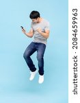 Portrait of young excited Asian man holding smart phone and jumping on isolated light blue studio background