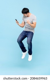 Portrait Of Young Excited Asian Man Holding Smart Phone And Jumping On Isolated Light Blue Studio Background