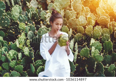 Portrait of young exquisite African American female drinking fresh coco water in front of wall of cactuses; cute black girl in chemise with coconut is slaking her thirst next to green hedge