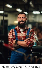 A portrait of a young factory worker with a tattoo on the forearm and a beard, wearing blue overalls and red and white plaid shirt. He is smiling with arms crossed. Factory setting.