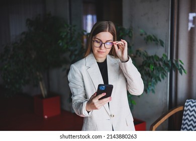 Portrait Of A Young Fair-haired Woman In A Beige Jacket And Glasses On Her Face, A Mod Phone In The Hands Of A Woman. Beautiful Modern Blogger Girl