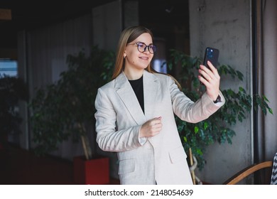 Portrait Of A Young Fair-haired Woman In A Beige Jacket And Glasses On Her Face, A Mod Phone In The Hands Of A Woman. Beautiful Modern Blogger Girl