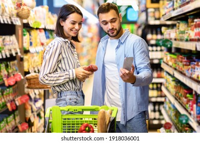 Portrait Of Young Family Doing Grocery Shopping Together In Supermarket. Smiling Couple Using Phone Scanning Food Product Standing In Store Aisles Indoors. Customers Buying Supplies, Comparing Price