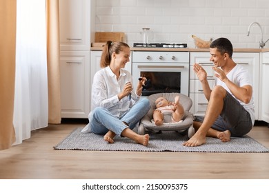 Portrait Of Young Family, Mom With Little Children Sit On Floor In New Modern Design Kitchen, Parents Playing With Their Baby In Rocking Chair And Showing Toys For Infant Daughter.