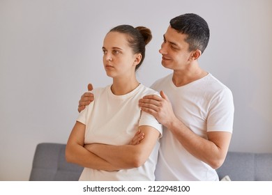 Portrait Of Young Family Wearing White Casual Style T Shirts Standing At Home In Room, Offended Wife Turning Away From Husband, Man Trying To Calm Down His Lovely Woman.