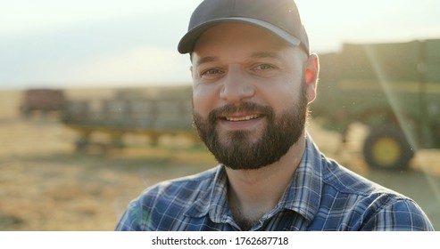 Portrait Of The Young Farmer In A Hat Looking At Combine Working In The Field With Corn And Then Turning His Head With A Smile To The Camera. Close Up. Rear.