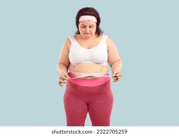 Portrait of a young fat overweight woman wearing sportswear measure the waist with measuring tape isolated on studio blue background. Obesity, weight control, sport and fitness concept.