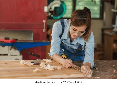 Portrait of young female Asian carpenter working in carpentry workshop,carpenter woman measuring wood plank on work bench, smiling Asian carpenter girl concentrate with her handicraft wood working job