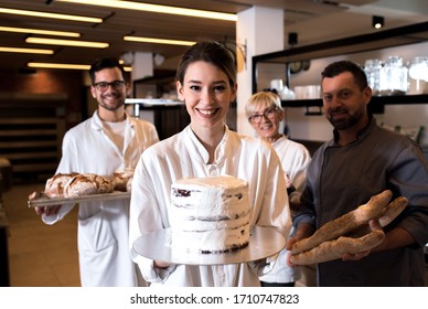 Portrait Of Young Female Baker Holding Unfinished Cake In Her Hands At Bakery With Her Multi Generation Colleagues In Backgrounds.