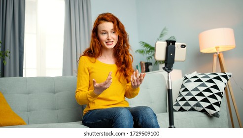 Portrait of young female blogger sitting on couch and talking in front of smartphone webcam. Girl coaching for her vlog online from home. Attractive happy woman coach talk for videoblog.