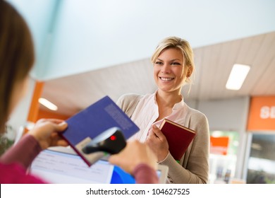 Portrait Of Young Female College Student Returning Book To Library, With Librarian Scanning Barcode