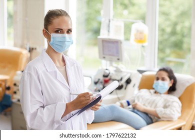 Portrait Of Young Female Doctor In Mask Writing In Medical Card And Looking At Camera With Patient In The Background