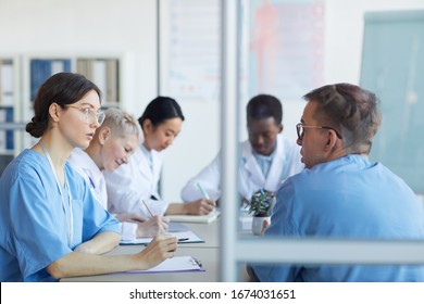 Portrait of young female doctor talking to colleague while sitting at table during medical council or conference in clinic, copy space