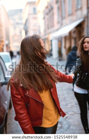 portrait of young female friends in colored leather biker jackets and hoodies holding hands while walking down the street. Stylish girls having fun, expressing positive emotions to camera.