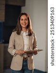 Portrait of young female leader standing at corporate office lobby with clipboard in hands and smiling at the camera. Smiling successful female executive at enterprise office with paperwork and docs