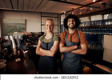 Portrait of young female and male staff wearing apron standing behind counter in cafe with crossed hands looking at camera - Powered by Shutterstock