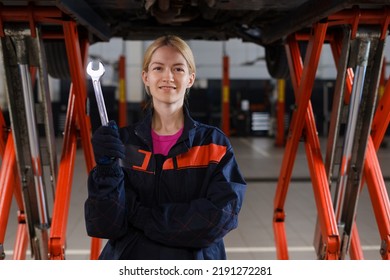 Portrait of a young female mechanic in uniform who holds a wrench and looks into the camera against the background of cars being repaired at a service station - Powered by Shutterstock