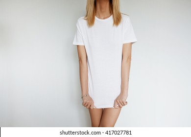 Portrait of young female model posing against home interior wearing only white blank copy space T-shirt while stretching it down. Trendy woman choosing clothes for going out to the cinema with friends