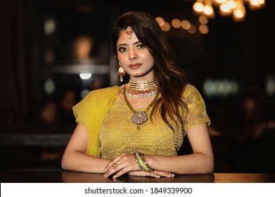 Portrait of Young female model sitting on sofa in a cafe, She is wearing Lehenga with heavy jewelry & makeup and Looking at camera with smile on face.