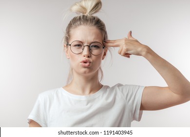 Portrait of young female student of european appearance making a suicide gesture, feeling bored and unhappy about coming session and exams, forming a gun with fingers, isolated against white wall