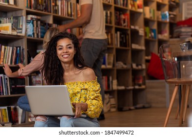 Portrait of young female student sitting at library using laptop with her friends in background.