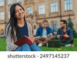 Portrait of young female student woman girl noting doing homework task sitting on the grass while two males college students classmates boys using laptop in background at university campus lawn