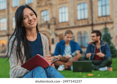 Portrait of young female student woman girl noting doing homework task sitting on the grass while two males college students classmates boys using laptop in background at university campus lawn - Powered by Shutterstock