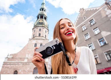 Portrait Of A Young Female Tourist With Photo Camera In Front Of Saint Peter Church In Riga. Woman Having Great Vacations In Latvia