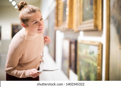 Portrait of young female visitor with guide book looking at exposition in art museum