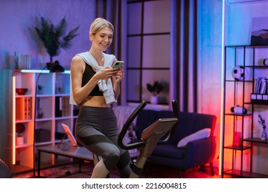 Portrait of young female wearing sport bra having conversation on her smartphone while working out using exercise bike. Home fitness workout sporty woman training on smart stationary bike indoors. - Powered by Shutterstock