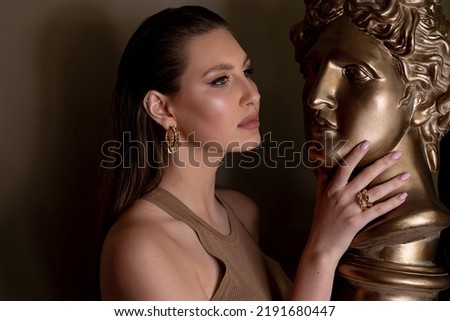 Portrait of young flawless thoughtful woman with long dark hair, bright golden make-up, pink lips touching head face of bronze bust David by Michelangelo on grey background. Beauty, cosmetics, art.
