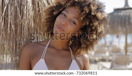 Portrait of young flirty girl with curls wearing white bikini and looking contently at camera while posing on resort beach in sunlight.