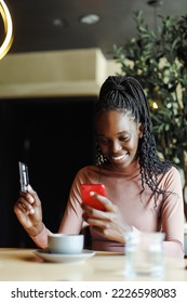 Portrait of young focused busy African-American woman with long dark braids wearing pink roll-neck sweater, sitting at table with cup of coffee in cafe, using red smartphone, giving credit card.  - Shutterstock ID 2226598083