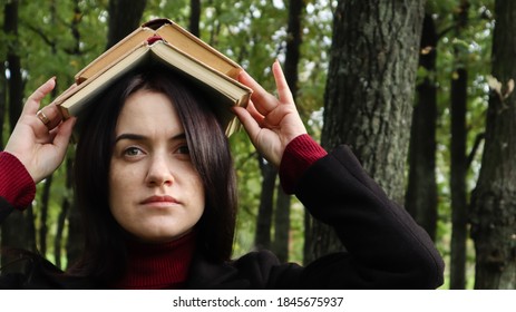 Portrait of a young and funny brunette in the park holding an open book on her head. Learning is fun. Woman balancing with books on her head. The student is tired of reading.