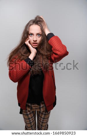 Portrait of a young funny girl with blue eyes and brown hair in a black T-shirt and a red bomber jacket. The girl holds on to her hair and fools around, has fun, grimaces, shows human emotions