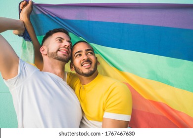 Portrait of young gay couple embracing and showing their love with rainbow flag at the street. LGBT and love concept.