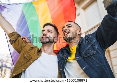 Portrait of young gay couple embracing and showing their love with rainbow flag at the street. LGBT and love concept.
