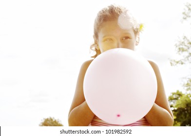 Portrait of a young girl blowing up a pink balloon in front of her face.