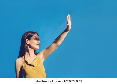 Portrait Of Young Girl Covering Her Face To Block Sun Light. Shadow And Light Over Woman's Face. Creative Portraits. Female In Orange Summer Beach Dress On A Blue Background