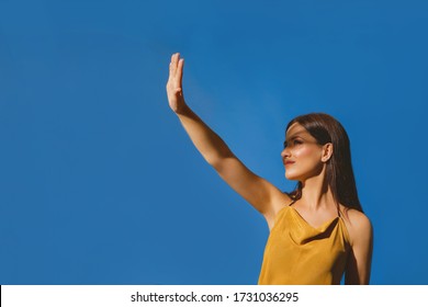 Portrait Of Young Girl Covering Her Face To Block Sun Light. Shadow And Light Over Woman's Face. Creative Portraits. Female In Orange Summer Beach Dress On A Blue Background