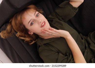 Portrait Of Young Girl In Dark Green Khaki Suit Laying On Black Sofa Barefoot. Pretty Blond European Girl With Short Hair-cut Smiling. Indoors. Doll Face Sexy Woman Relax At Home